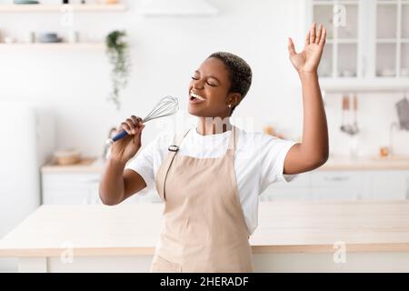 Felice ispirazione giovane afroamericana in grembiule prepara il cibo, canta al microfono immaginario Foto Stock