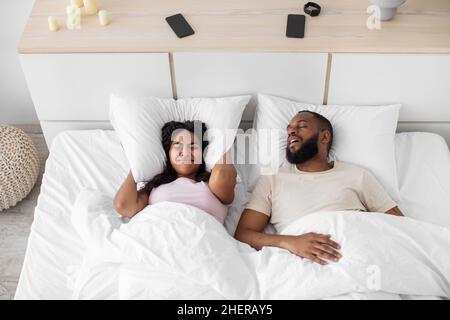 Arrabbiato giovane moglie afroamericana copre le orecchie con cuscino e soffre di rumore del marito Foto Stock