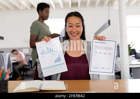Ritratto di una donna d'affari creativa asiatica sorridente che mostra le carte mentre si siede alla scrivania in ufficio Foto Stock
