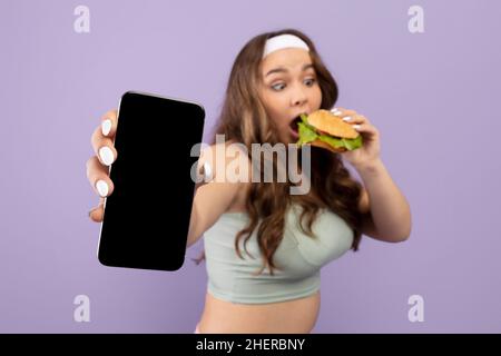 Giovane donna caucasica affamata e divertente più taglia in sport uniforme mangiare hamburger e smartphone show con schermo vuoto Foto Stock