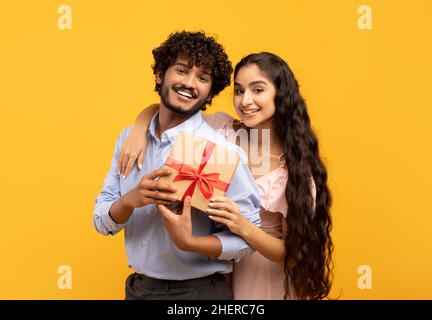 Ritratto di amorevole coppia indiana che tiene avvolto il regalo per San Valentino insieme, posando su sfondo giallo studio Foto Stock