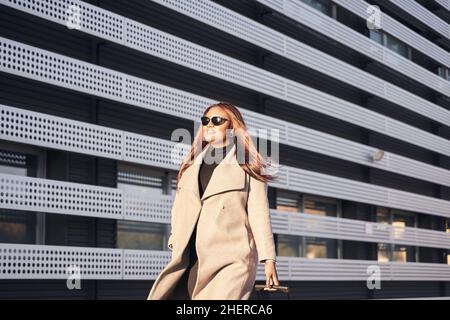 Ritratto di donna afro-americana con occhiali da sole e cappotto beige che cammina con valigia in mano fuori dalla stazione. Foto Stock
