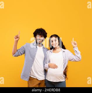 Eccitata signora incinta indiana e suo marito che punta verso l'alto allo spazio libero, mostrando vendita o offerta stupefacente, sfondo giallo Foto Stock