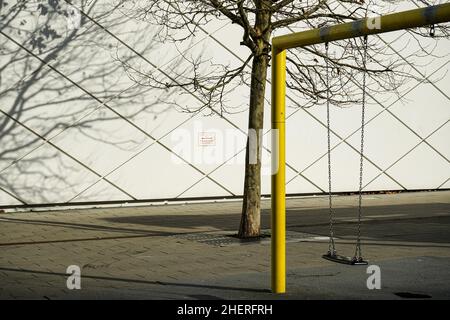 Un'altalena da gioco per bambini in un parco giochi. Un albero proietta ombre su un muro di casa. Foto Stock