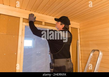 Costruzione di sauna, rifinitura. L'uomo sta avvitando una panca di legno al muro. Foto Stock