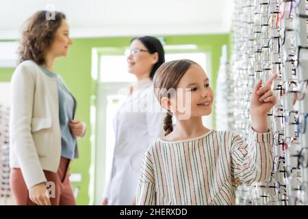 sorridente ragazza che punta a assortimento di occhiali vicino mom blurred e oculist asiatico in negozio di ottica Foto Stock