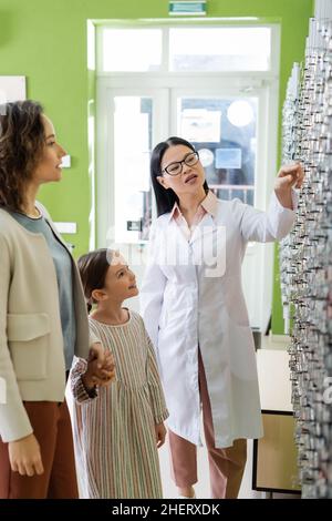oftalmologo asiatico che punta agli occhiali vicino a madre e figlia in negozio di ottica Foto Stock