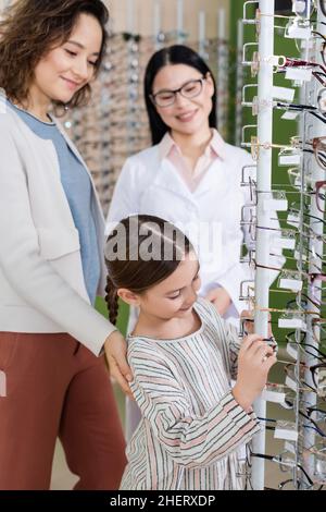 bambino che sceglie gli occhiali vicino a mamma sorridente e offuscato oculista asiatico in negozio di ottica Foto Stock