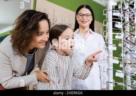 ragazza stupita che punta agli occhiali vicino a mamma sorridente e offuscato oculista asiatico in ottica salone Foto Stock