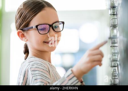 sorridente ragazza in occhiali puntando con il dito in ottica negozio in primo piano sfocato Foto Stock