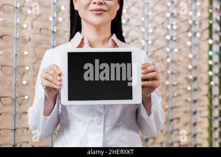 vista ritagliata dell'oculista sfocato in cappotto bianco che tiene il laptop con schermo vuoto nel salone dell'ottica Foto Stock