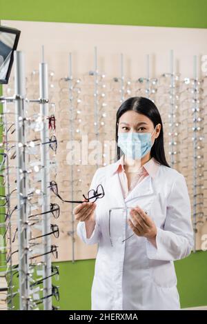 oculista asiatico in maschera medica confrontando occhiali mentre si lavora in negozio di ottica Foto Stock