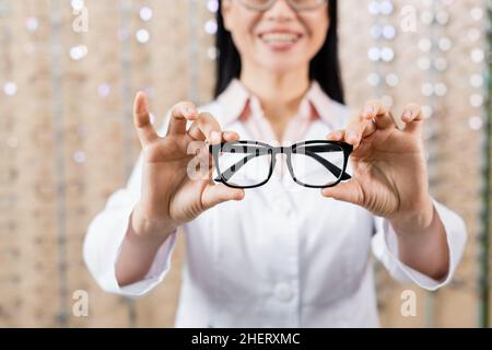 vista parziale dell'oculista che mostra gli occhiali mentre sorride su sfondo sfocato Foto Stock