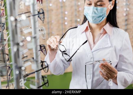 oftalmologo asiatico in maschera medica confrontando occhiali in negozio di ottica Foto Stock