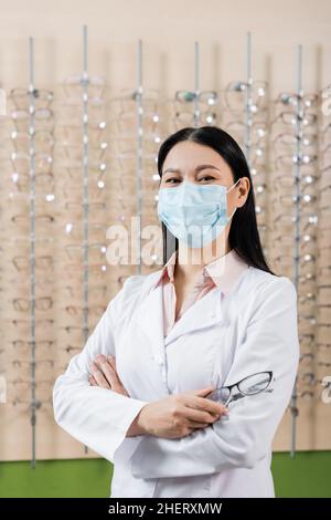 oftalmologo asiatico in maschera medica in piedi con braccia incrociate vicino offuscato assortimento di occhiali in negozio di ottica Foto Stock