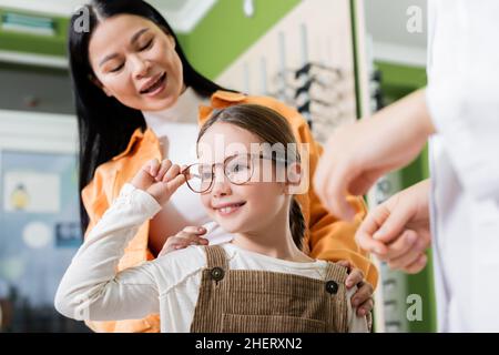 ragazza felice che regola gli occhiali vicino sorridente mamma asiatica e sfocato oculista in negozio di ottica Foto Stock