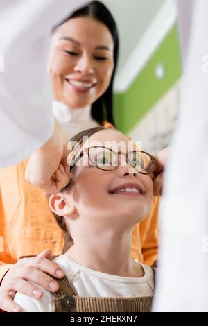 offuscato oculista cercando occhiali su ragazza allegra vicino donna asiatica in negozio di ottica Foto Stock