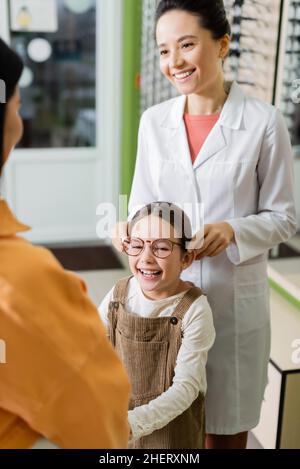 oculista positivo cercando occhiali sulla ragazza allegra vicino alla madre sfocata in negozio di ottica Foto Stock