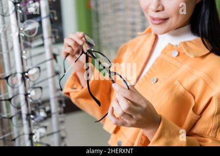 vista ritagliata della donna sfocata confrontando occhiali in negozio di ottica Foto Stock