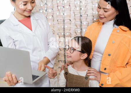 ragazza sorridente in occhiali puntando al portatile vicino oculista e madre asiatica in ottica salone Foto Stock