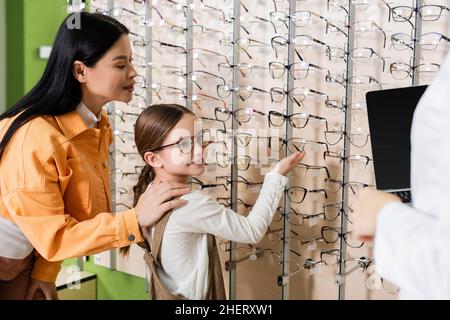 ragazza sorridente che punta a assortimento di occhiali vicino a mamma asiatica e oculista sfocato con laptop in negozio di ottica Foto Stock