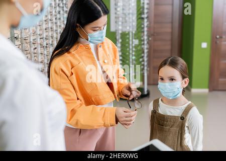 donna asiatica in maschera medica che tiene gli occhiali vicino alla figlia e offuscato oculista in negozio di ottica Foto Stock