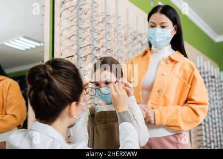 oftalmologo blurred che prova gli occhiali sulla ragazza nella maschera medica vicino alla donna asiatica nel deposito dell'ottica Foto Stock
