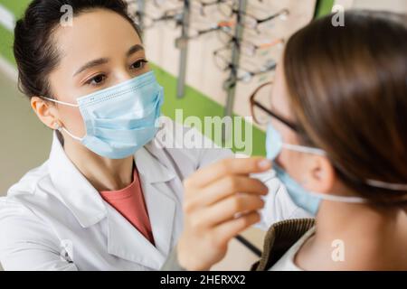 oculista in maschera medica cercando occhiali sulla ragazza sfocata in negozio di ottica Foto Stock