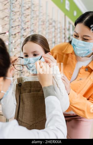 offuscato oculista cercando occhiali sulla ragazza in maschera medica vicino asiatico madre in ottica salone Foto Stock