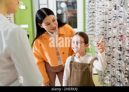 positivo capretto che sceglie gli occhiali vicino al mom asiatico e oftalmologista sfocato in negozio di ottica Foto Stock
