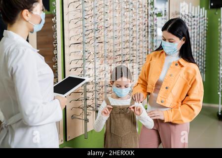 ragazza in maschera medica scelta occhiali vicino asiatico madre e sfocato oculista con tavoletta digitale Foto Stock