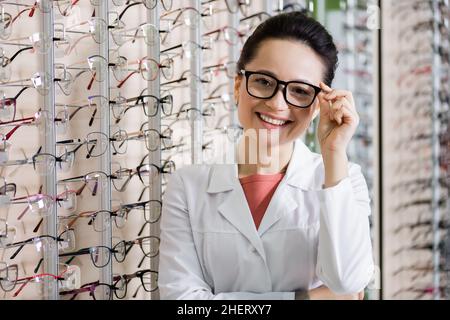 felice oftalmologo che guarda la macchina fotografica mentre regola gli occhiali in negozio di ottica Foto Stock
