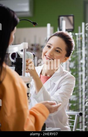 giovane optometristo sorridente vicino donna sfocata e oftalmoscopio in negozio di ottica Foto Stock