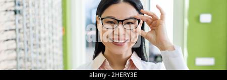 gioioso oculista asiatico regolare occhiali mentre si guarda la fotocamera in ottica salone, banner Foto Stock