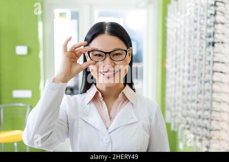 felice oculista asiatico regolare occhiali mentre si guarda la fotocamera in negozio di ottica Foto Stock