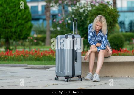 Triste giovane donna bionda in pantaloncini, una camicia è seduta con una valigia su una panca contro lo sfondo di alberi, cespugli in una giornata estiva Foto Stock