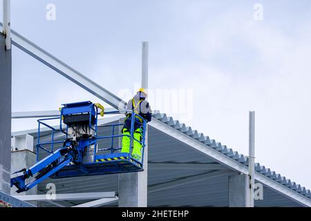 un operatore che utilizza un ascensore semovente in materiale uniforme e di sicurezza monta le strutture metalliche di un edificio Foto Stock