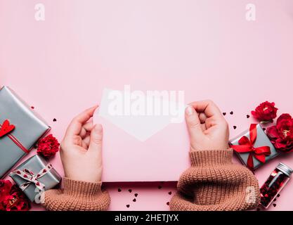 Le mani femminili tengono il messaggio vuoto della lettera di amore nella busta aperta, il biglietto di auguri di San Valentino o il mock in bianco di invito in su. Spazio di copia, vista dall'alto. Sfondo rosa con cuori, scatole regalo grigie, confetti Foto Stock