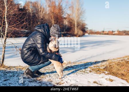 Donna che cammina pug cane nel parco nevoso invernale da lago ghiacciato abbraccio animale domestico. Buon cucciolo che indossa l'imbracatura in piedi sulle gambe posteriori. Foto Stock