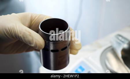 Primo piano delle mani in guanti medici che mettono polvere chimica dal cucchiaio per sostanze chimiche al piccolo contenitore di metallo in laboratorio. Mani inserimento del reagente bianco Foto Stock