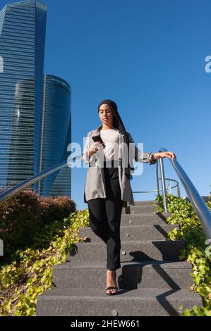 Immagine verticale di una donna adulta che indossa un foulard nero che scende su una scala pubblica in città utilizzando il telefono con edifici dietro in una d soleggiato Foto Stock