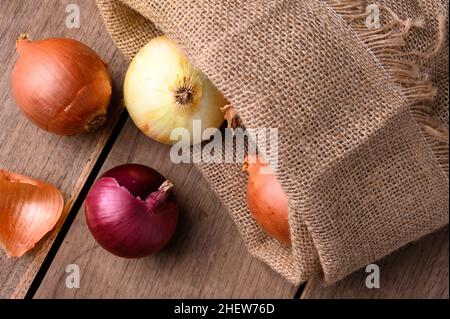 Cipolle rosse e bianche su un rustico tavolo di legno che fuoriesce da un sacco di stoffa in cucina, vista dall'alto. Foto di alta qualità Foto Stock