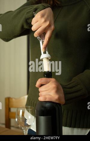 Primo piano su una donna in una giacca verde che rimuove il sughero da una bottiglia di vino rosso su un tavolo Foto Stock