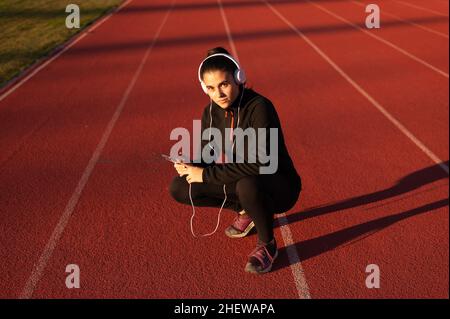 la giovane atleta guarda il cellulare con le cuffie sulla pista di atletica Foto Stock