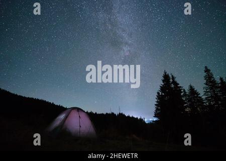 Luminosa tenda turistica illuminata che risplenderà sul campeggio in montagne scure sotto il cielo notturno con stelle scintillanti. Concetto di stile di vita attivo. Foto Stock