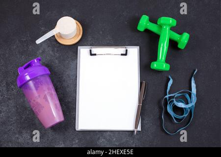 Agitatore e misurino di polvere proteica, manubri, nastro di misurazione e notebook su tavolo scuro. Allenamento e dieta fitness. Concetto di stile di vita sano. Foto Stock