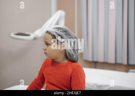 Ritratto a profilo di bambina in cappuccio medico monouso e maglia rossa in attesa di procedure mediche in ospedale o dentale ufficio. Foto Stock