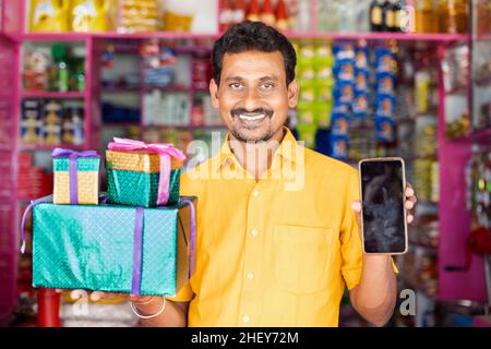 Kirana Shop Merchant con scatole regalo e telefono cellulare in mano loking alla macchina fotografica - concPET di vendite Festival, offerte promozionali per vincere premi da Foto Stock
