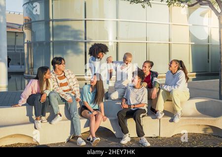 Grande gruppo di amici adolescenti felici che parlano in una panchina nella strada del campus studentesco. Foto Stock