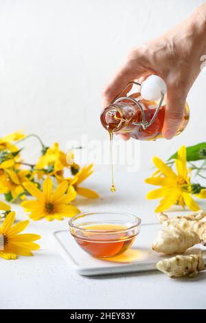 Versare lo sciroppo di carciofo di Gerusalemme in una ciotola, fiori e radici su sfondo bianco. Formato verticale. Sostituto dello zucchero per dolci vegani crudi. swe naturale Foto Stock